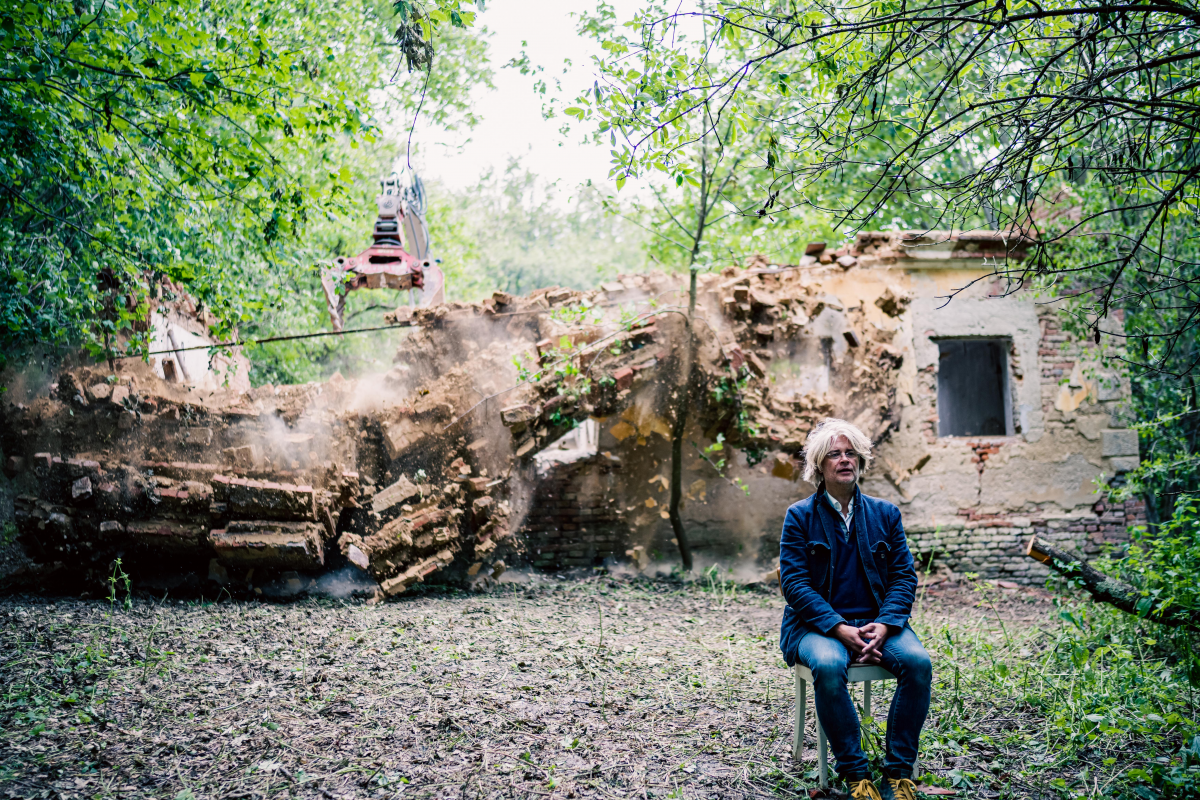 Edgar Honetschläger sitzt rechts im Bild auf einem Stuhl im Wald. Hinter ihm ist ein Gebäude zu sehen das abgerissen wird.