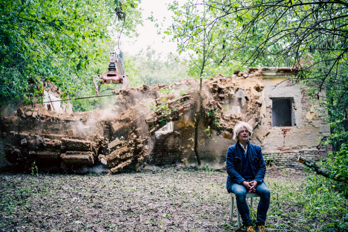 Edgar Honetschläger sitzt rechts im Bild auf einem Stuhl im Wald. Hinter ihm ist ein Gebäude zu sehen das abgerissen wird.